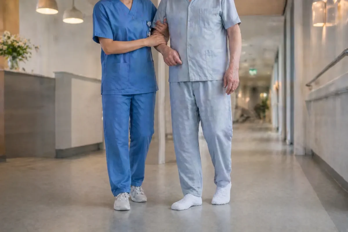 Nurse assisting an elderly patient walking in a hospital ward corridor, patient wearing white non-slip grip socks visible at foot level