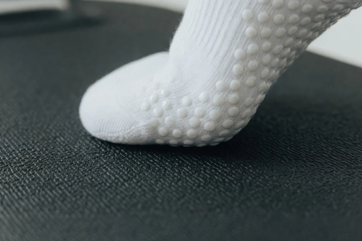 Close-up detail of a white full-toe grip sock pressed against the textured non-slip surface of a Megaformer carriage platform, showing silicone grip dots making contact